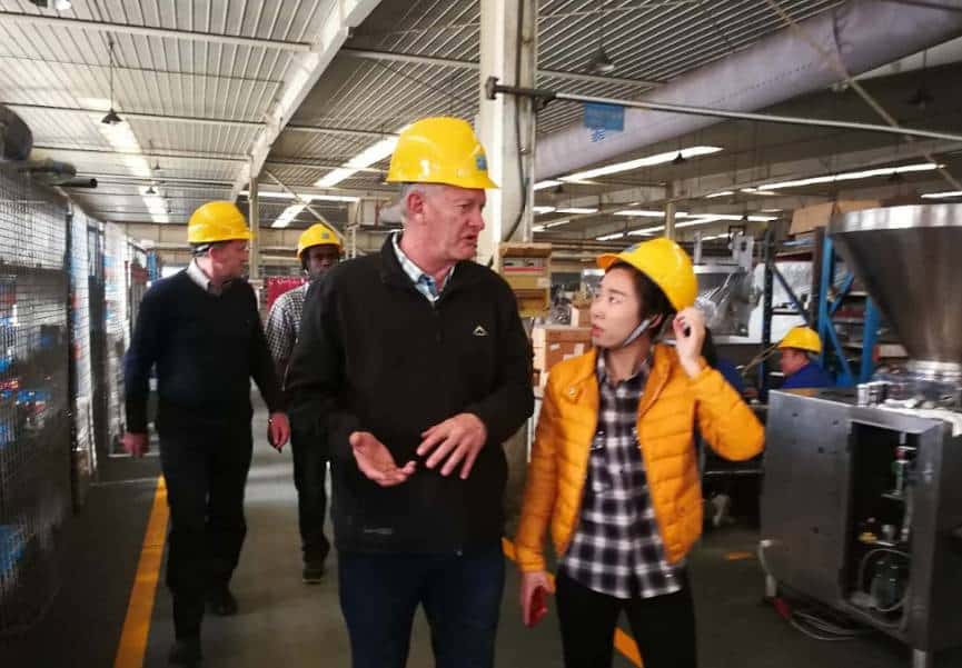 a group of people wearing yellow hard hats inside an industrial or manufacturing facility, with a man in a black jacket talking to a woman in a yellow jacket while others walk behind them