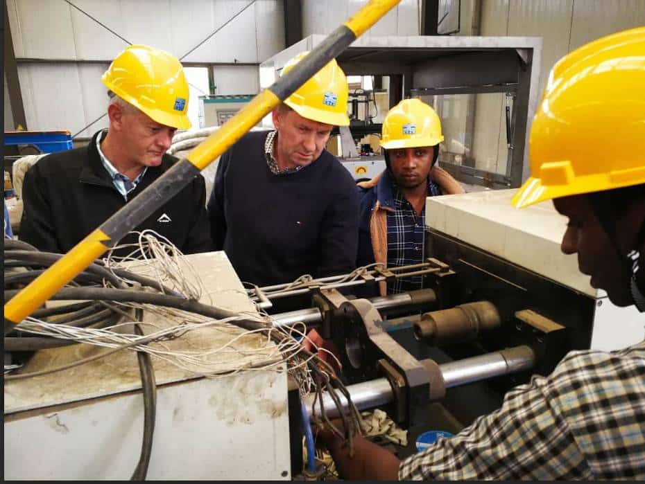  a group of individuals wearing yellow hard hats observing and inspecting a piece of industrial machinery in a factory setting