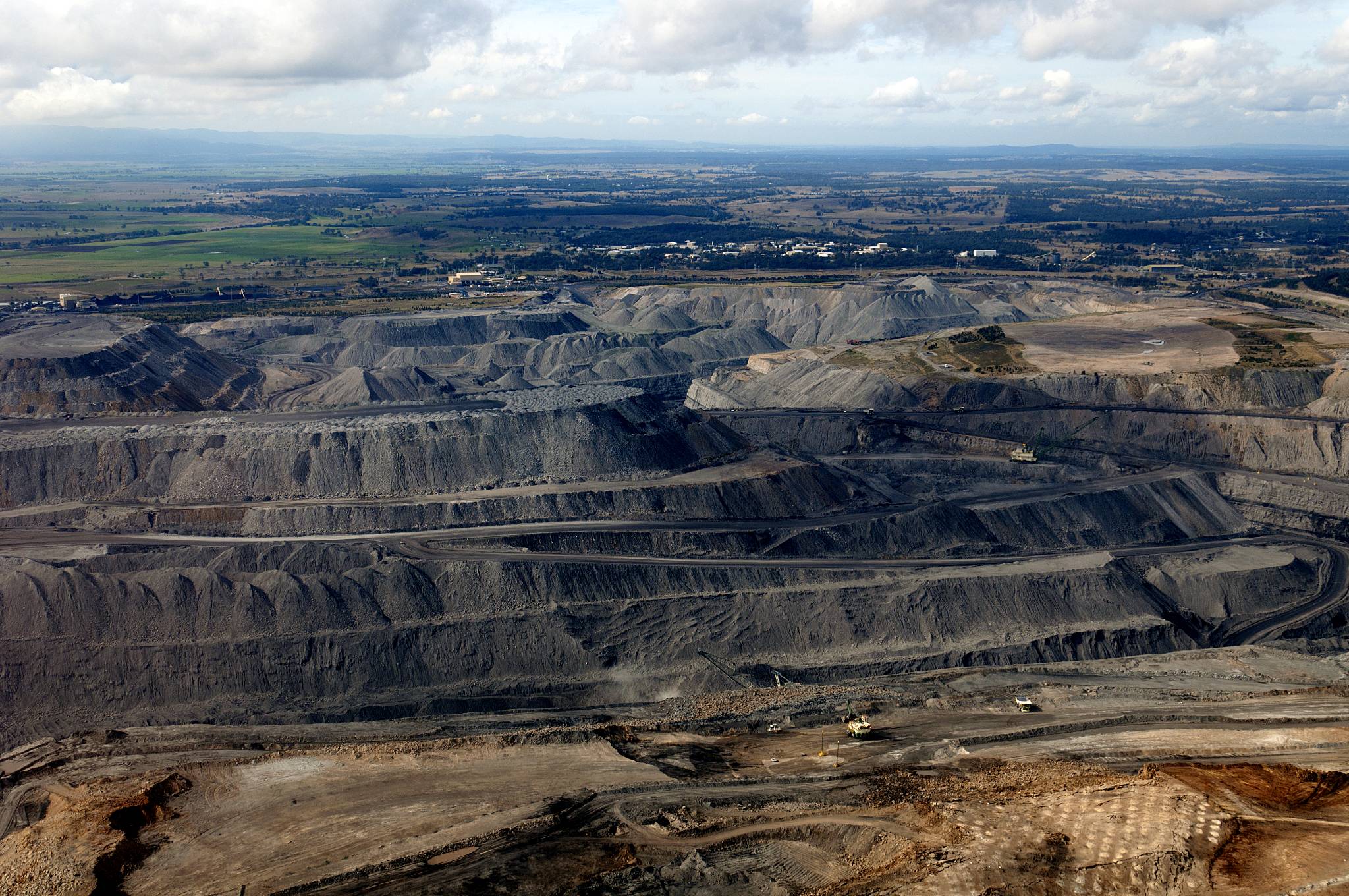 Aerial view of a large open-pit mining operation with multiple terraced levels carved into the earth, surrounded by green plains in the distance.