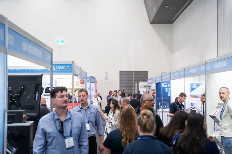 Attendees walking through a crowded exhibition aisle lined with branded booths at a WA Mining trade show