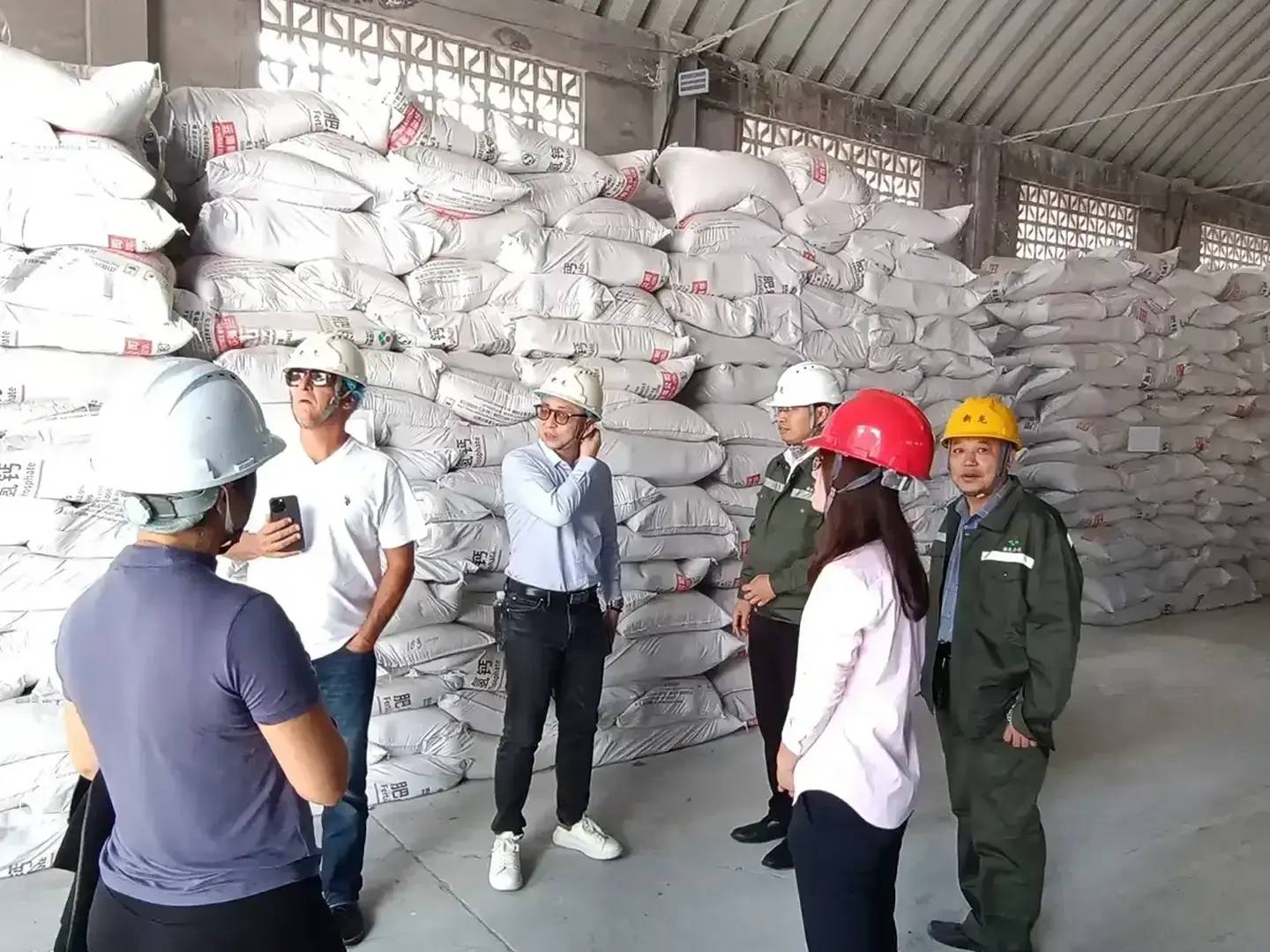 A group in hard hats inspecting stacked sacks of industrial chemical product inside a Chinese factory warehouse