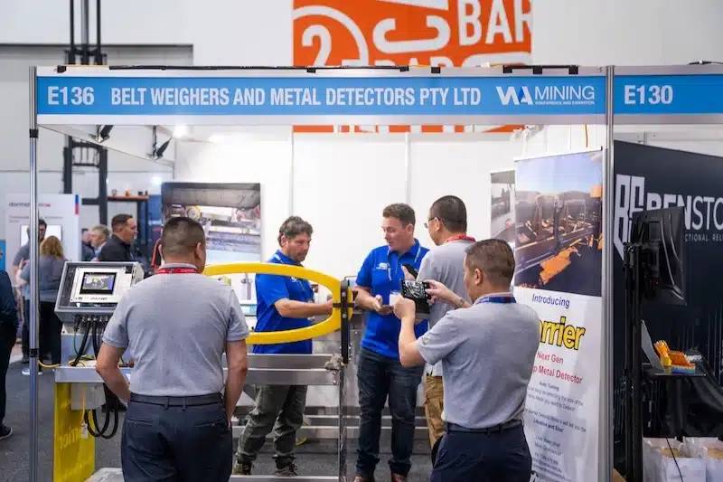Exhibitors demonstrating a belt weigher and metal detector to a small group of attendees at a branded trade show booth