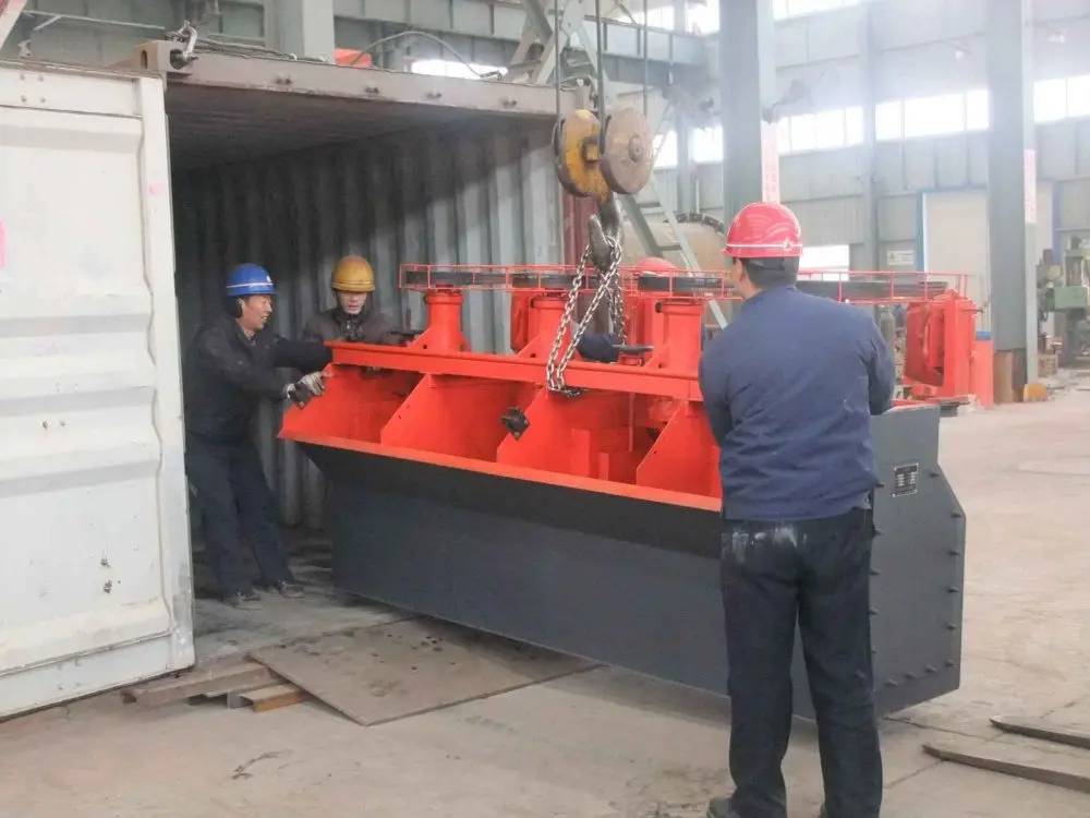 Workers using a crane hook to load an orange flotation processing unit into a shipping container at a factory loading bay